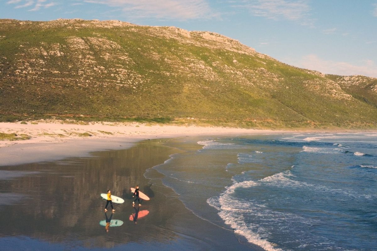 Two surfers walking into the ocean with Cape Town mountain scenery in the background.